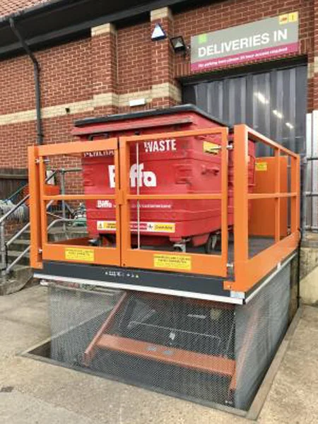 An orange loading bay lift situated in a loading area, with a large red bin inside it